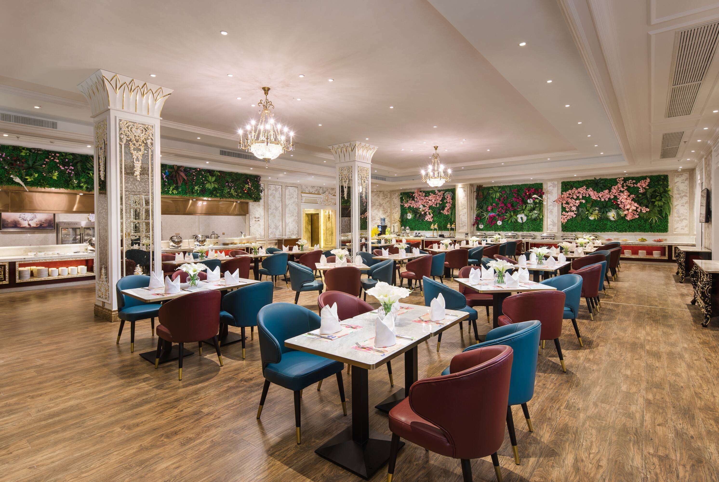 A spacious restaurant with multiple dining tables and chairs, elegant chandeliers, and decorative wall panels.