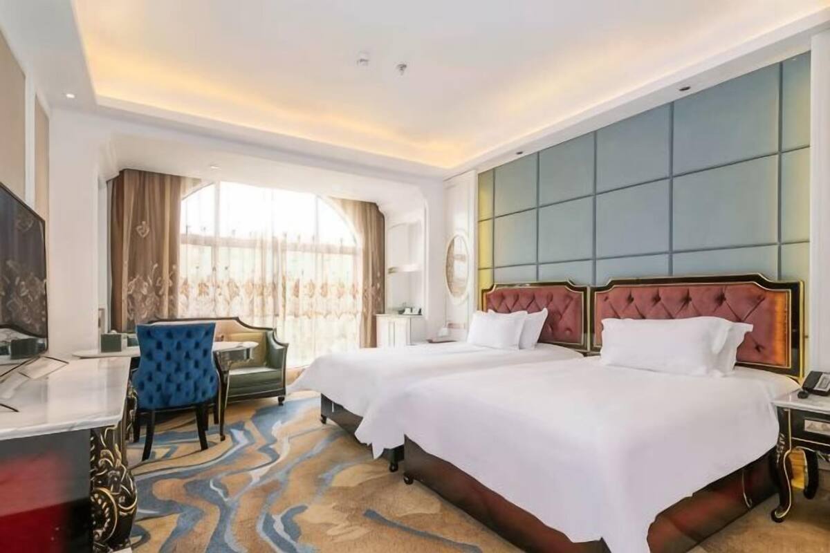A hotel room with two beds, a desk, a chair, a TV, and a large window with curtains.