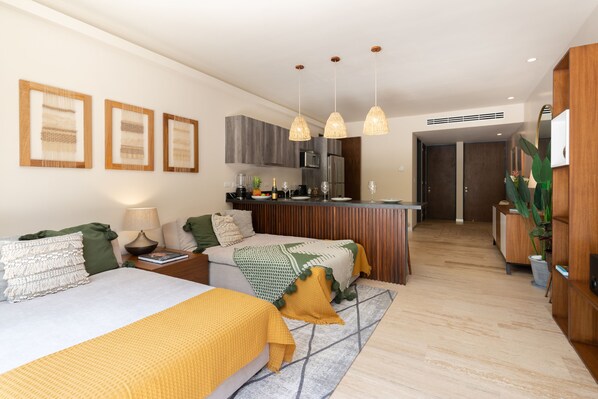 A modern hotel room with a sofa, two beds, a kitchen area, and a hallway leading to a door. - Casa Luna 1BR by the Pool - Attha Cenote (Tulum)