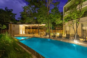 A swimming pool with lounge chairs and a covered seating area, surrounded by trees and a building with large windows. - Casa Luna 1BR by the Pool - Attha Cenote (Tulum)