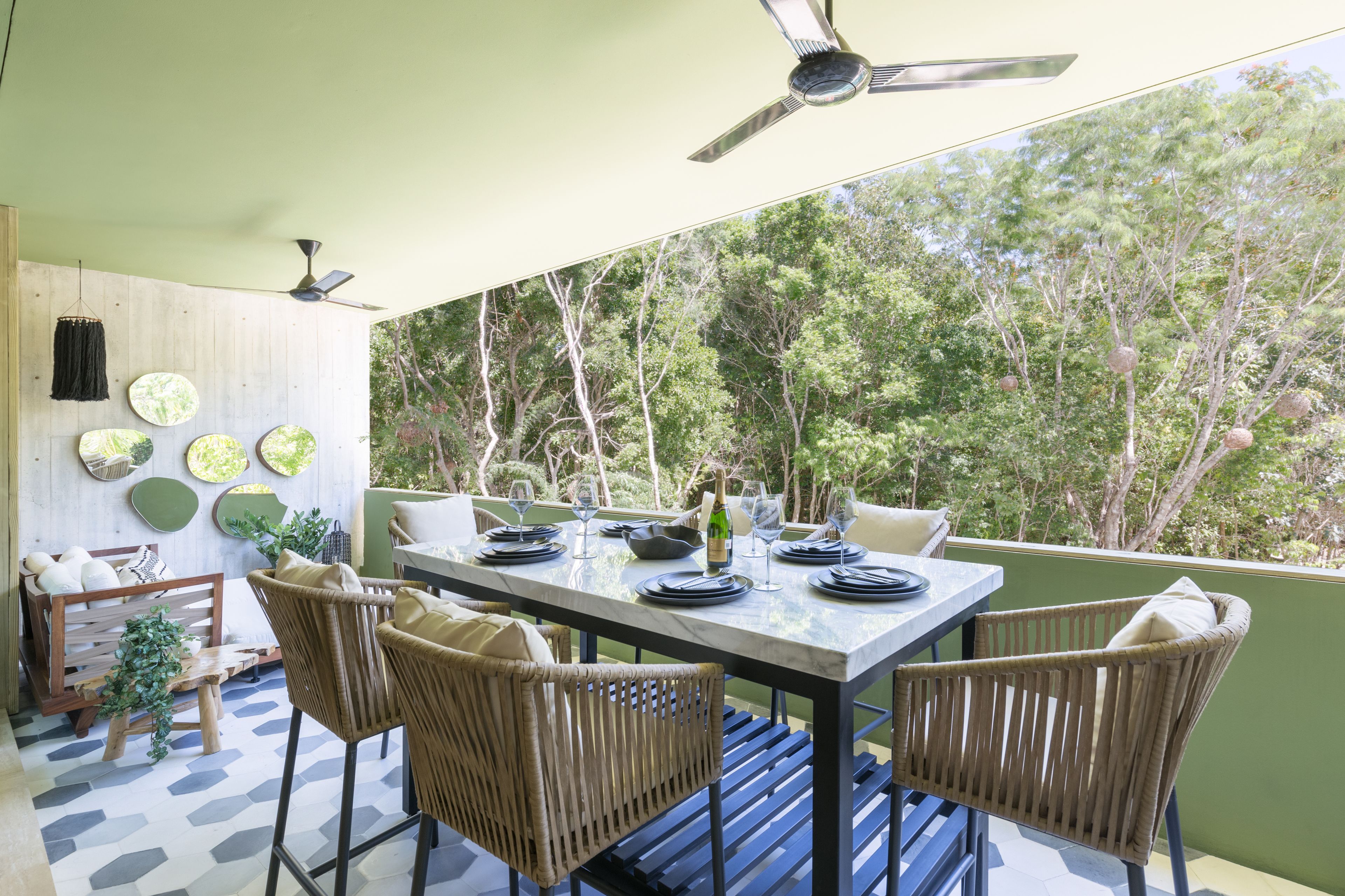 A dining area with a table set for six, surrounded by wicker chairs, a ceiling fan, and a view of trees.