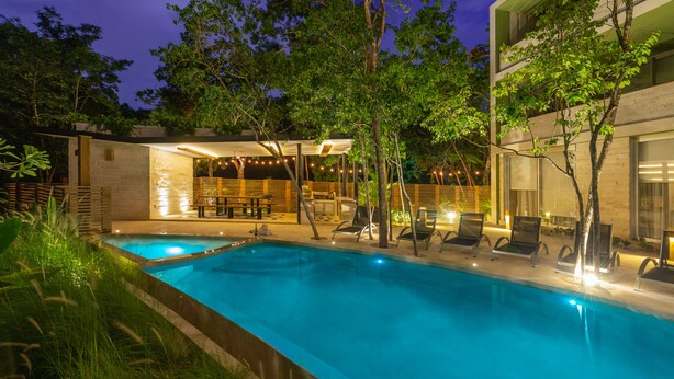 Una piscina con sillones de descanso y un área de estar techada, rodeada de árboles y un edificio con grandes ventanales.