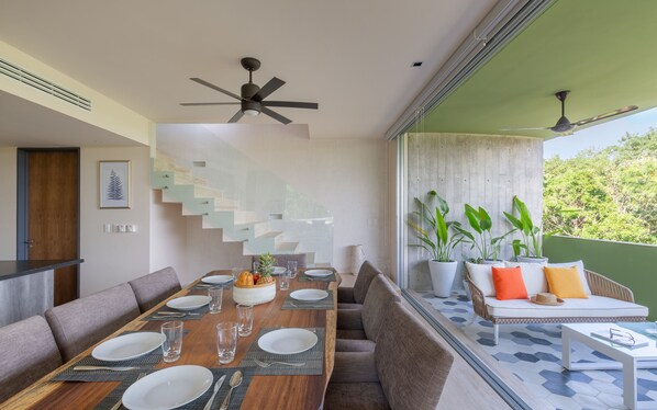 A dining area with a wooden table set for four, a staircase with a glass railing, and a seating area with a sofa and plants.