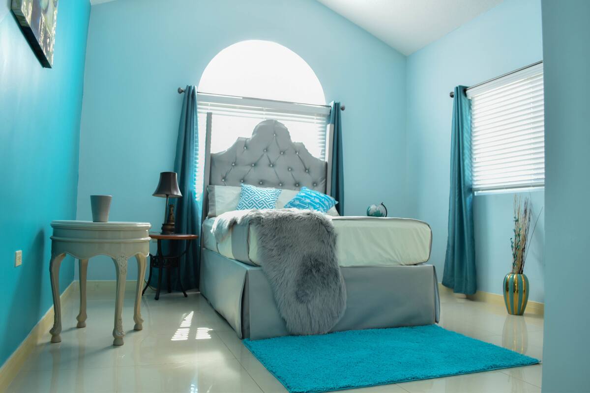 Une chambre à coucher avec un lit, une table de chevet, une lampe, un vase et une fenêtre avec des rideaux.