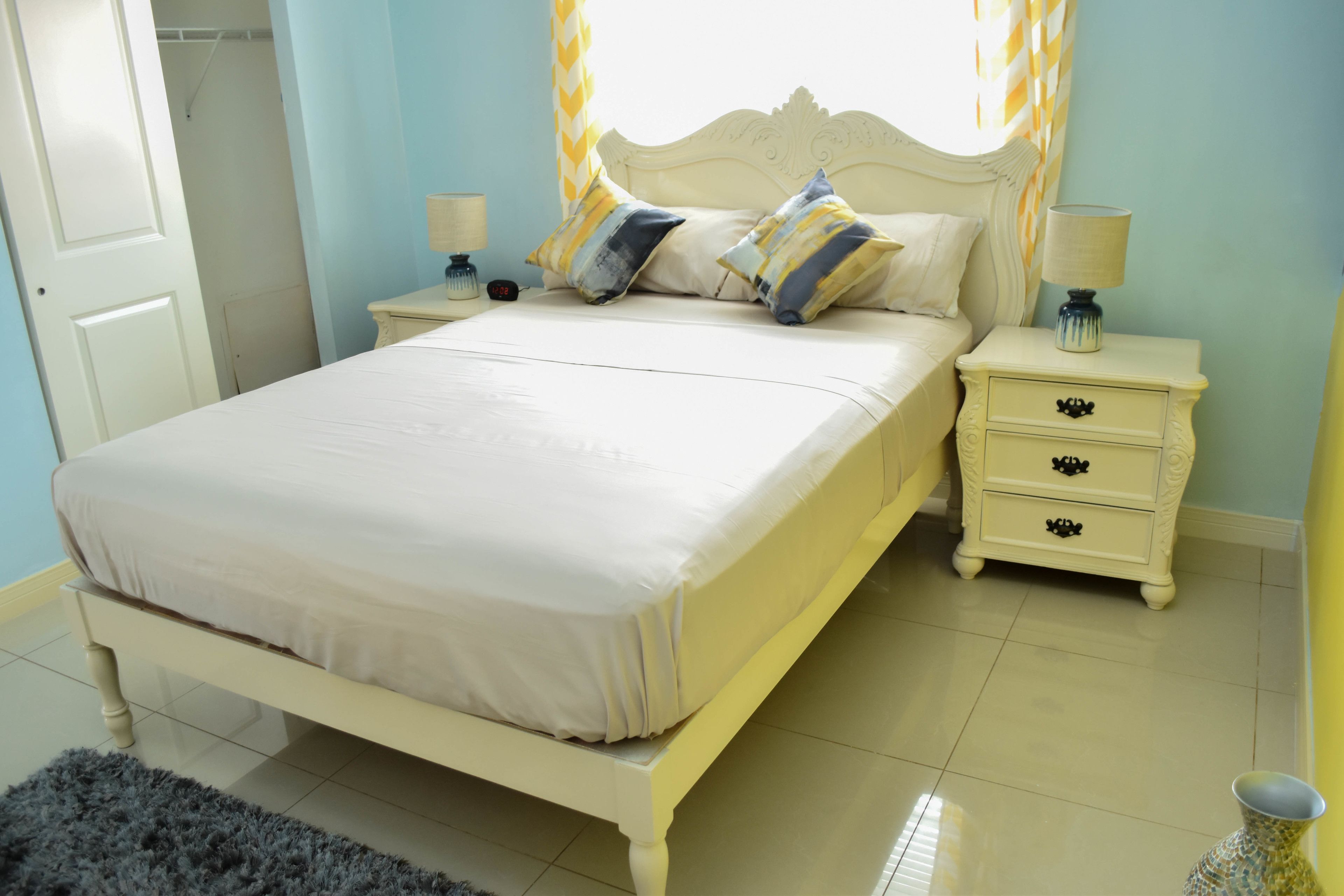 Un lit recouvert d’une couvre-lit blanche et agrémenté de deux oreillers, une table de chevet avec une lampe et une petite table de nuit avec un vase.