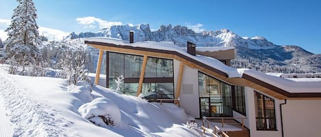 A modern chalet with large glass windows, a wooden deck, and a view of snow-covered mountains.