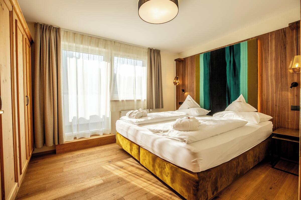 A neatly made bed with white linens and towels, a wooden floor, and a large window with sheer curtains.
