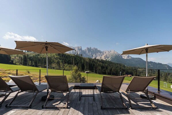 A terrace with four lounge chairs and two umbrellas, overlooking a mountainous landscape.