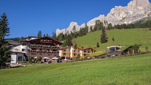 A hotel with multiple balconies adorned with flowers, situated in a green valley with mountains in the background. - Moseralm Dolomiti Hideaway (Nova Levante)