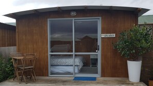 Une cabane en bois avec une porte coulissante en verre, un lit à l’intérieur, une petite table avec des chaises, et une plante en pot à l’extérieur.