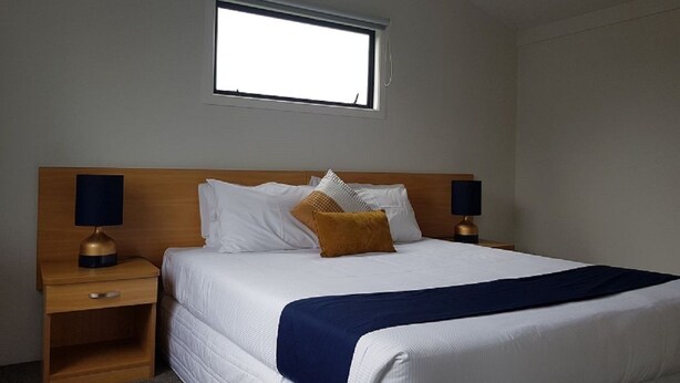 Un lit bien fait, recouvert d’une couverture bleue, avec deux lampes de chevet et une fenêtre au-dessus du lit.