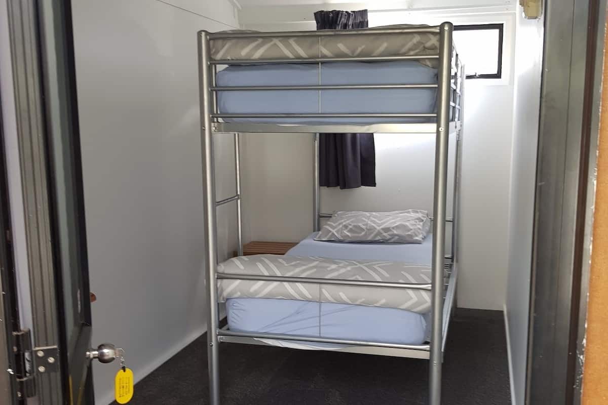 Ein Etagenbett mit Metallgestell, einer Matratze und einem Kopfkissen. Eine Tür mit Griff und einem Schlüsselbund sind ebenfalls zu sehen.