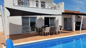 Un espace aménagé au bord de la piscine, comprenant une table et des chaises, un patio couvert et un bâtiment doté d’un balcon.