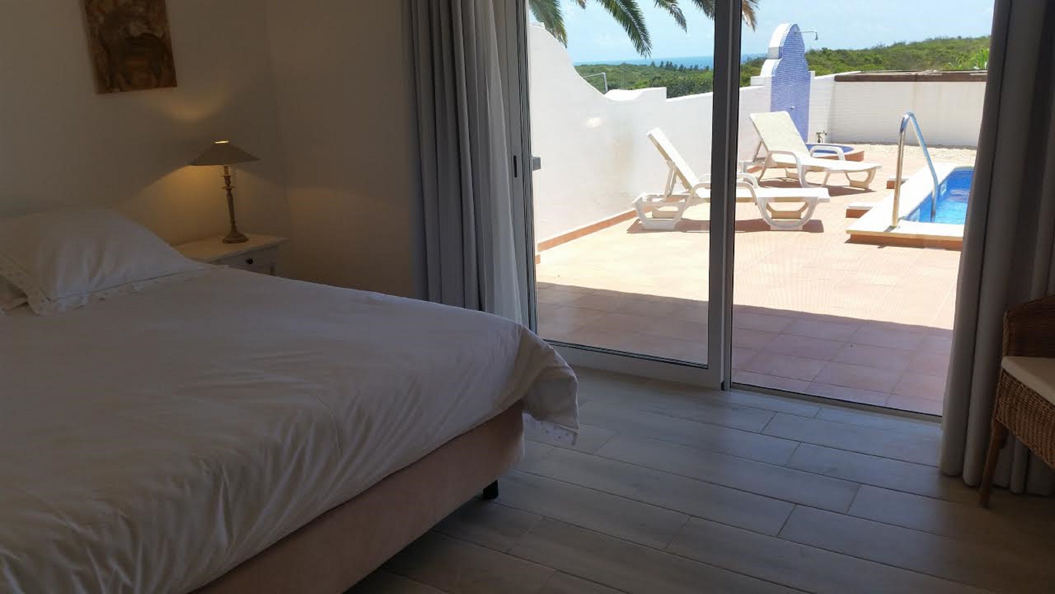 Un dormitorio con cama, mesita de noche, lámpara y vista a una piscina y sillas de descanso a través de una puerta corrediza de vidrio.