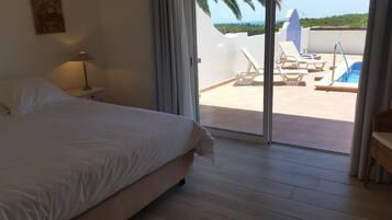 Un dormitorio con cama, mesita de noche, lámpara y vista a una piscina y sillas de descanso a través de una puerta corrediza de vidrio.