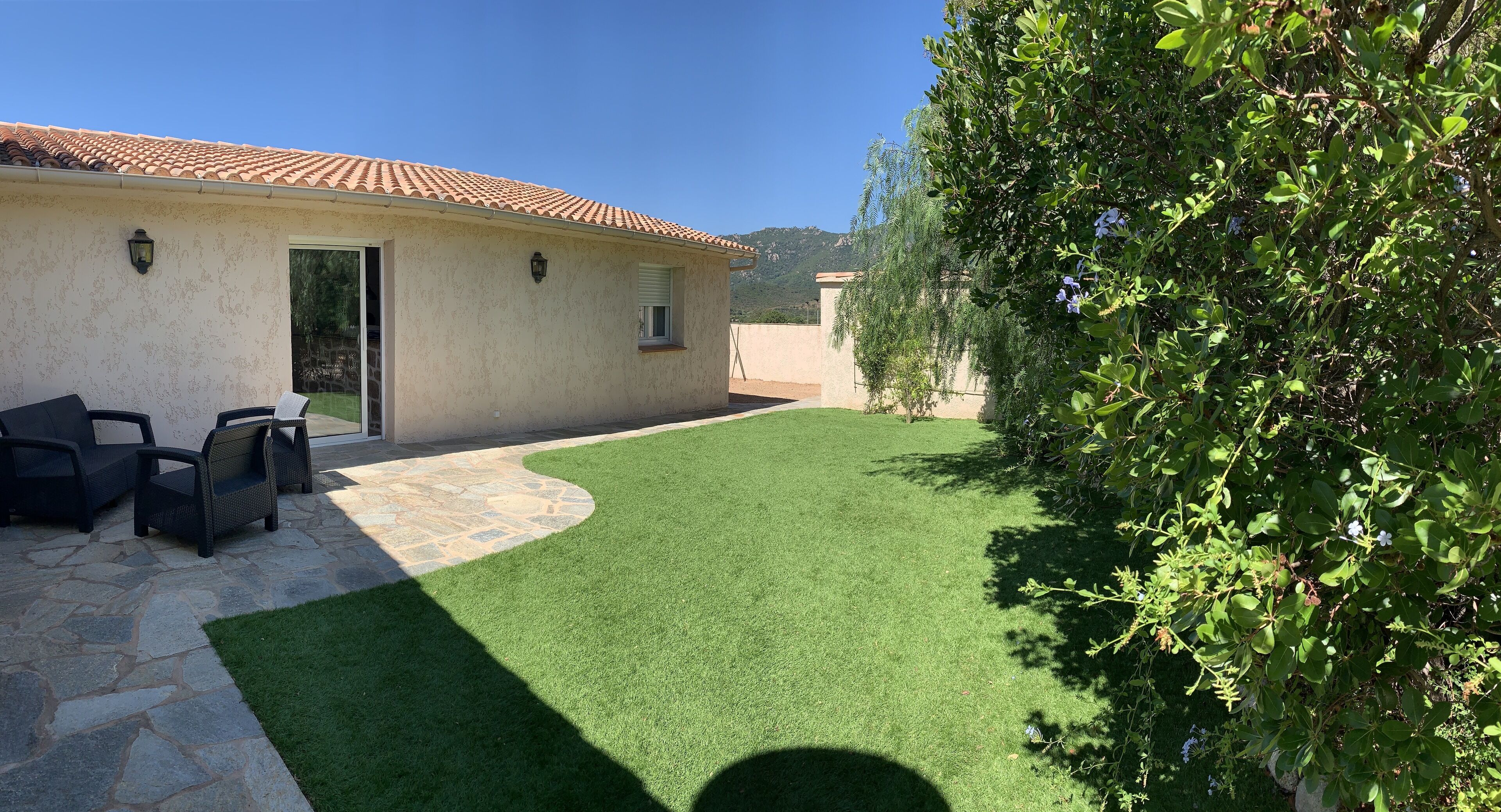 A patio area with a stone floor, a small seating area with chairs, and a green lawn.