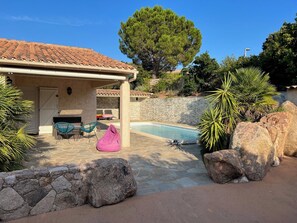 Un espace piscine doté d’un patio en pierre, de chaises longues et d’un coin salon abrité.