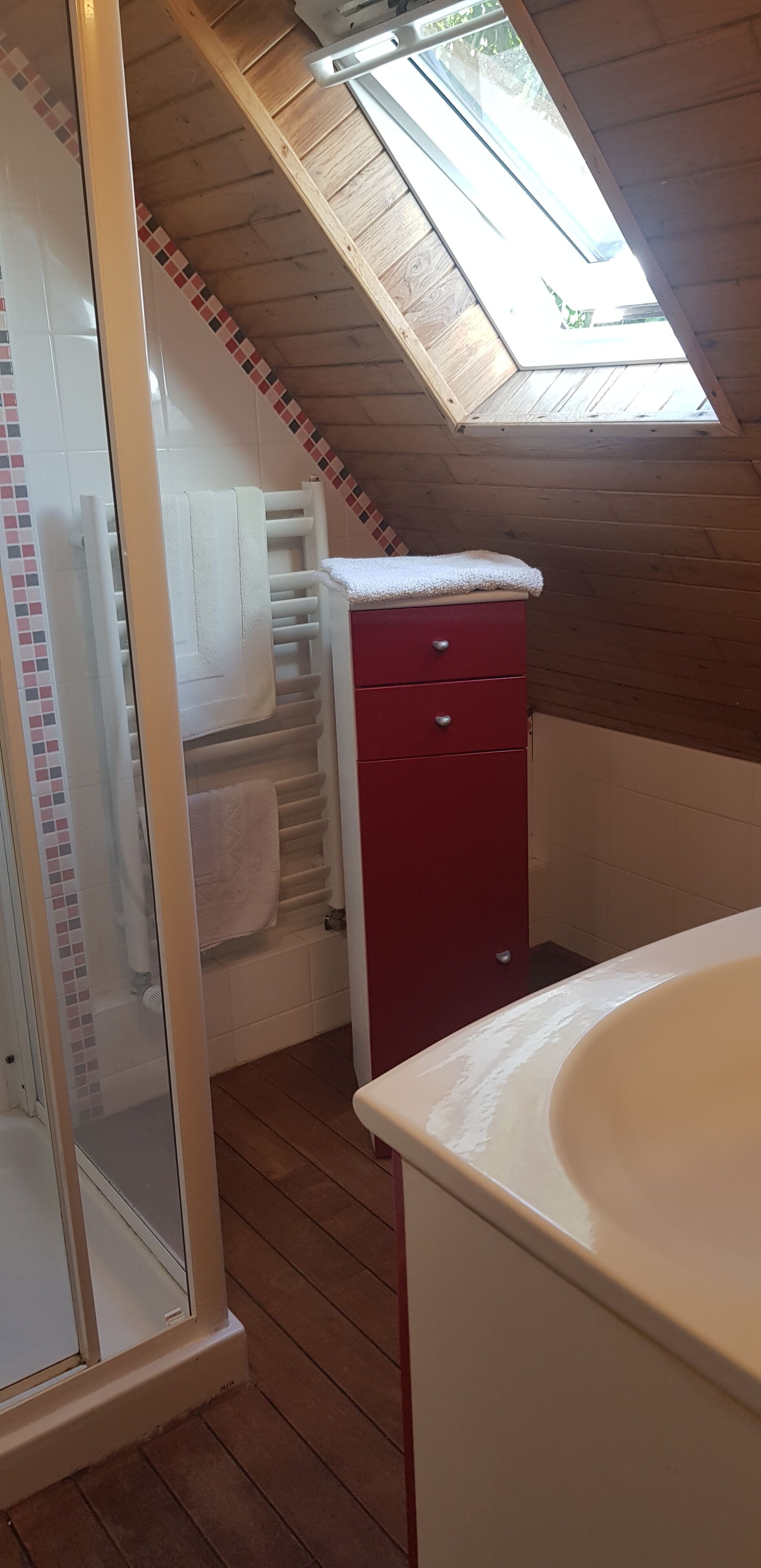 Un baño pequeño con tragaluz, ducha, un armario rojo y un lavamanos blanco.