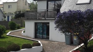 Une maison moderne avec un toit en pente, un balcon et un jardin agrémenté d’un arbre et d’un parterre de fleurs.