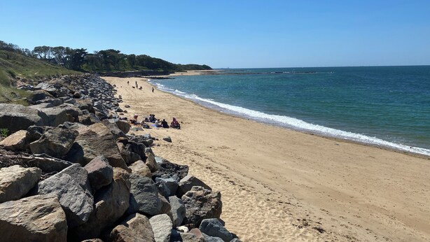 A beach with large rocks in the foreground, a sandy shore, and a clear blue sky.