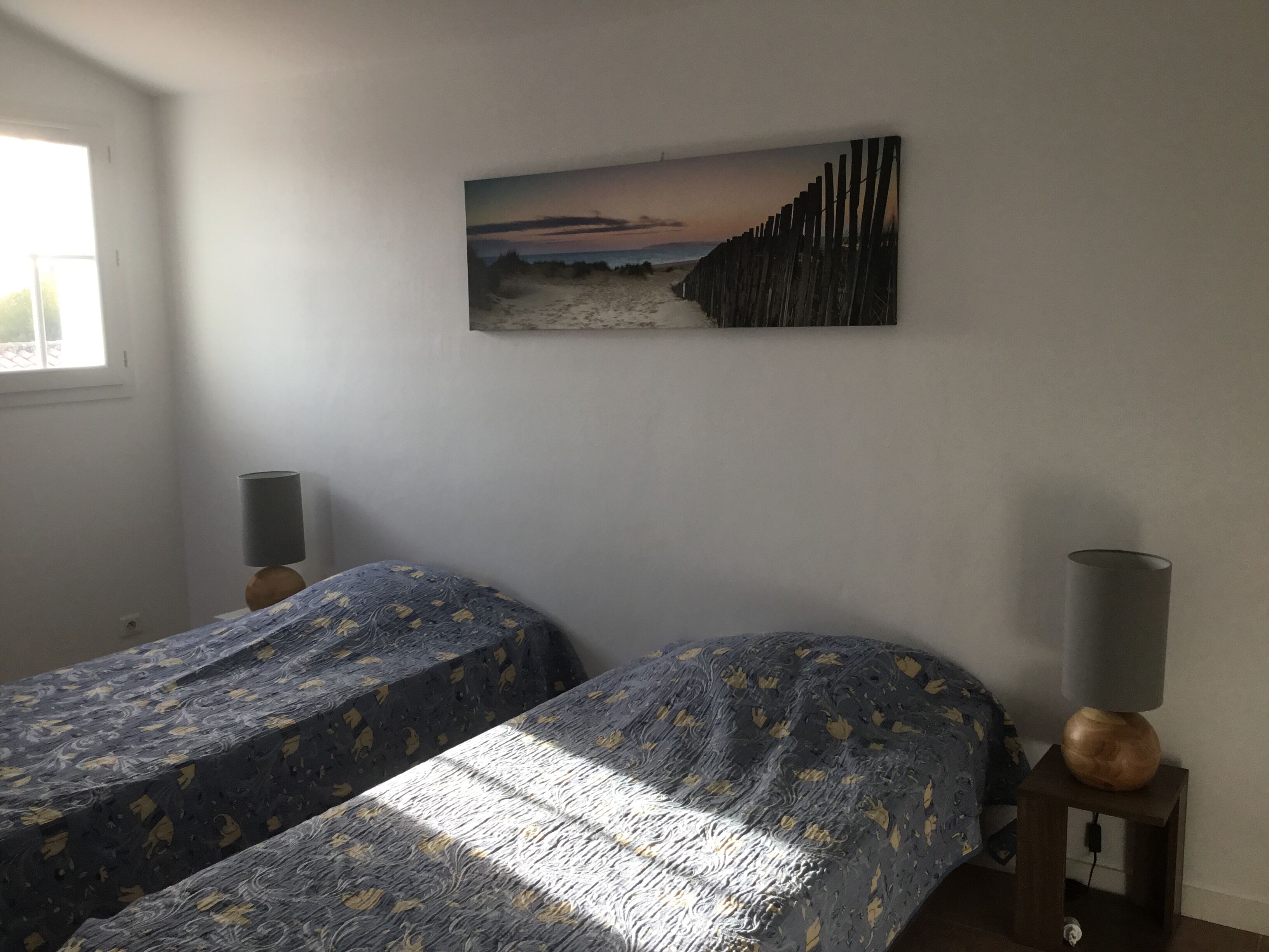 A bedroom with two beds, a painting of a beach scene, and a window providing natural light.