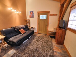 A cozy living area with a black leather sofa, a patterned throw pillow, a small wooden side table, a television on a wooden stand, and a door with a window above it.