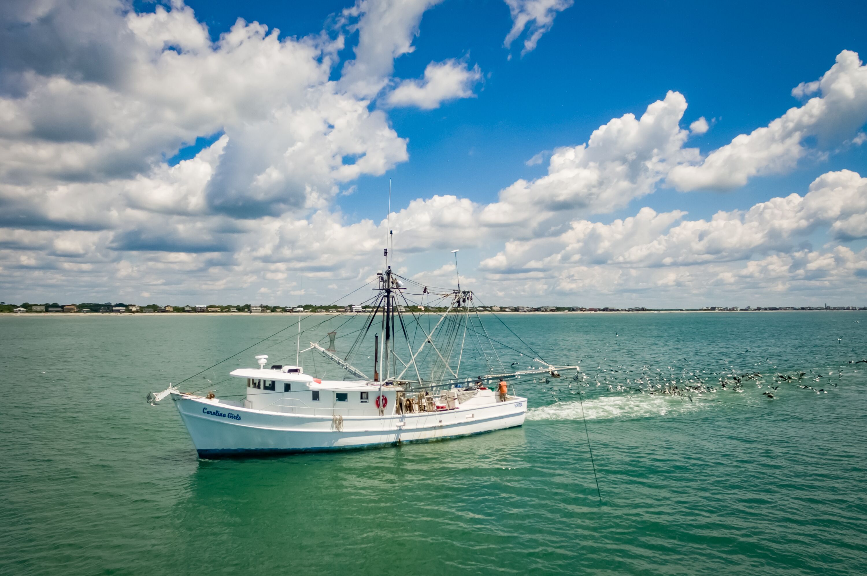 A boat named 'Carolina Girls' on calm turquoise waters with a clear sky and scattered clouds.