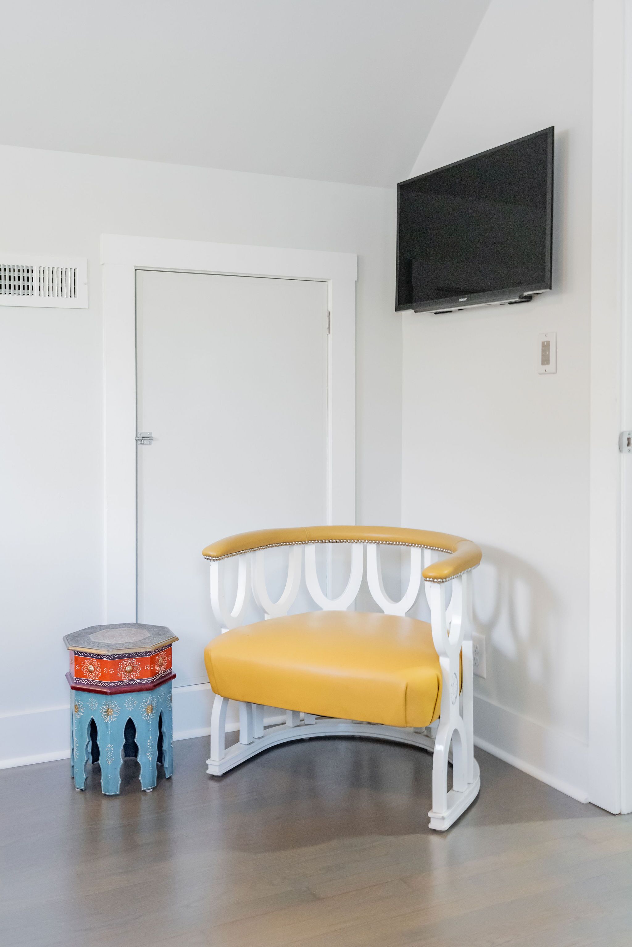 A room with a white door, a television mounted on the wall, a yellow cushioned bench, and a small blue stool with decorative patterns.