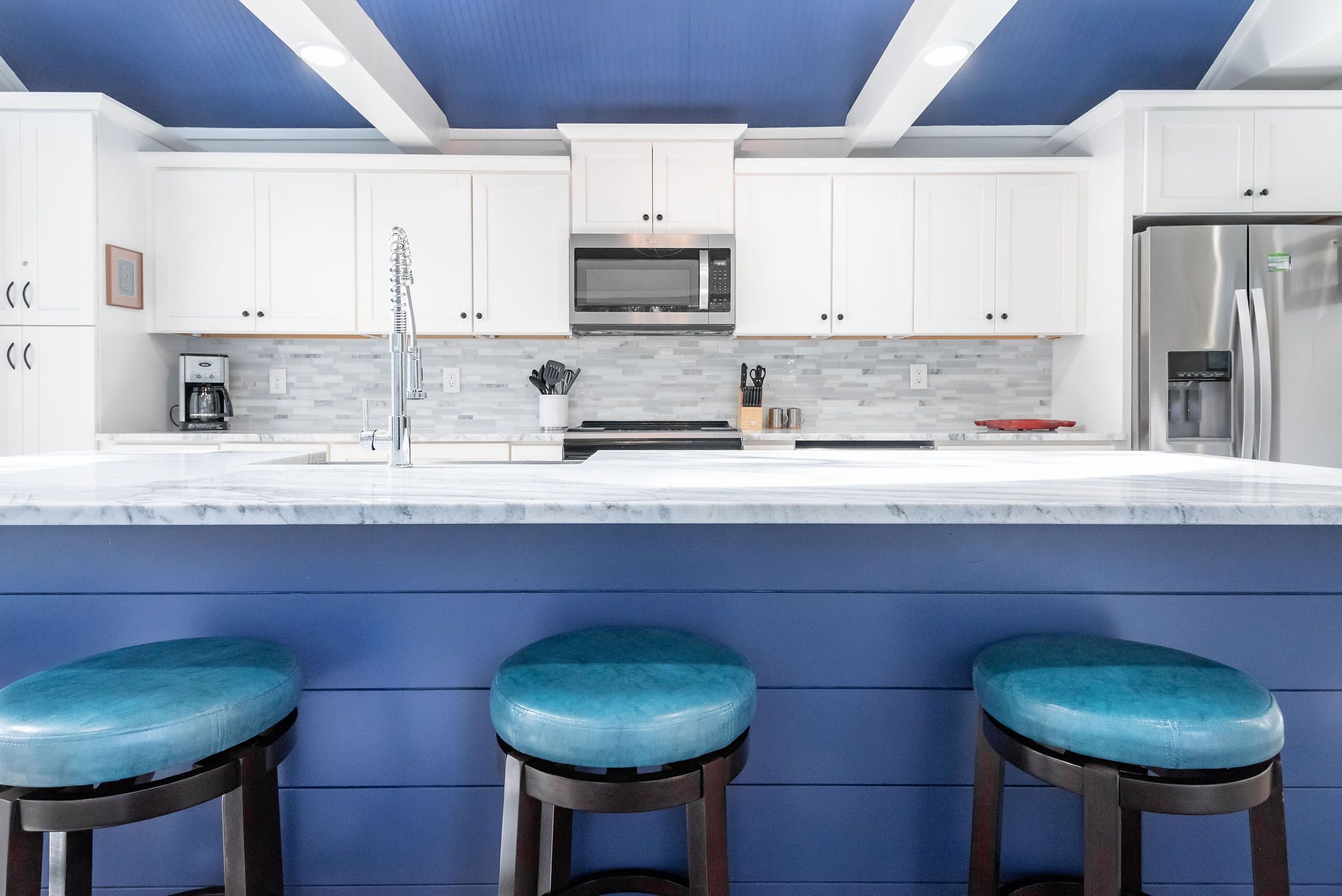 Eine moderne Küche mit weißen Schränken, einem Edelstahlkühlschrank, einer Mikrowelle und einer Kaffeemaschine. Drei blaue Barhocker stehen an der Arbeitsplatte.