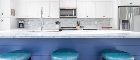 Eine moderne Küche mit weißen Schränken, einem Edelstahlkühlschrank, einer Mikrowelle und einer Kaffeemaschine. Drei blaue Barhocker stehen an der Arbeitsplatte.