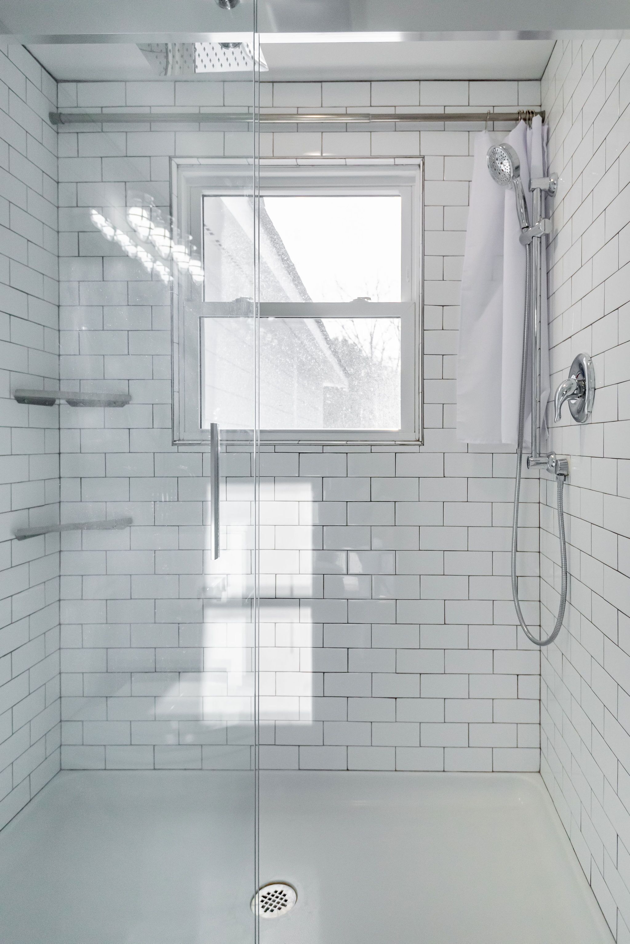 A modern bathroom with white tiled walls, a glass shower enclosure, a window, and a handheld showerhead.