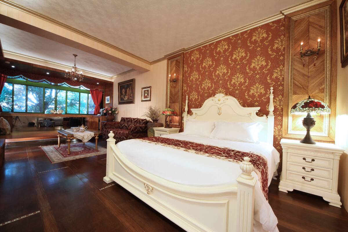 A spacious bedroom with a large bed, a nightstand, a dresser, and a window with a view of trees.