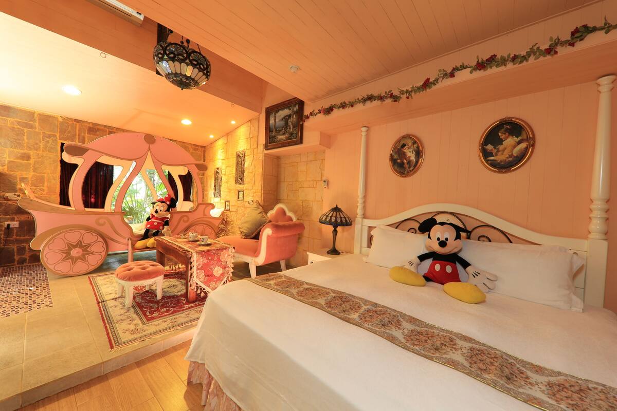 A bedroom with a large bed, Mickey Mouse plush toy, a stone wall, and a chandelier.