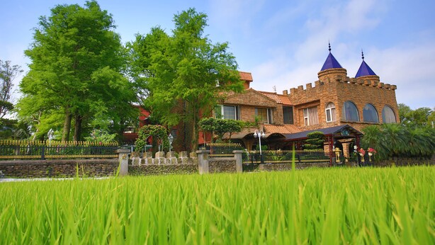 Un edificio de estilo castillo con fachada de piedra y dos cúpulas azules, rodeado de exuberante vegetación y un jardín cuidado.