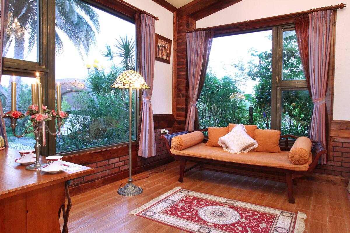 A cozy room with a wooden floor, a sofa with cushions, a table with a lamp, and a window with curtains.