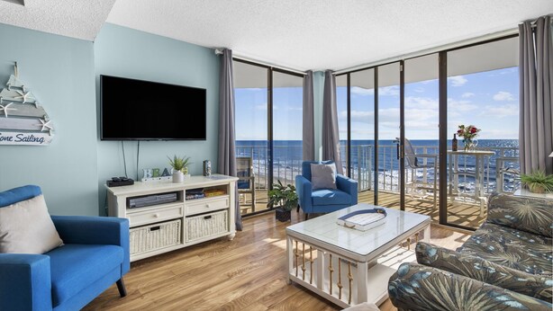 A modern living room with a large TV, a blue sofa, a white coffee table, and a view of the ocean through floor-to-ceiling windows. - Beautiful Ocean Front 5th Flr Condo (North Myrtle Beach)