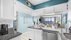 A kitchen with white cabinets, granite countertops, and a dishwasher. There is a coffee maker on the counter, and a window with a view of the city. - Beautiful Ocean Front 5th Flr Condo (North Myrtle Beach)