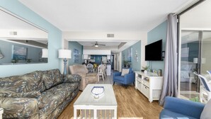 A living room with a sofa, a coffee table, and a TV. There is a kitchen in the background with a dining table and chairs. - Beautiful Ocean Front 5th Flr Condo (North Myrtle Beach)