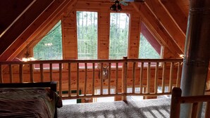 Eine Holzhütte mit Balkon und Geländer, durch große Fenster blickt man auf einen Wald.