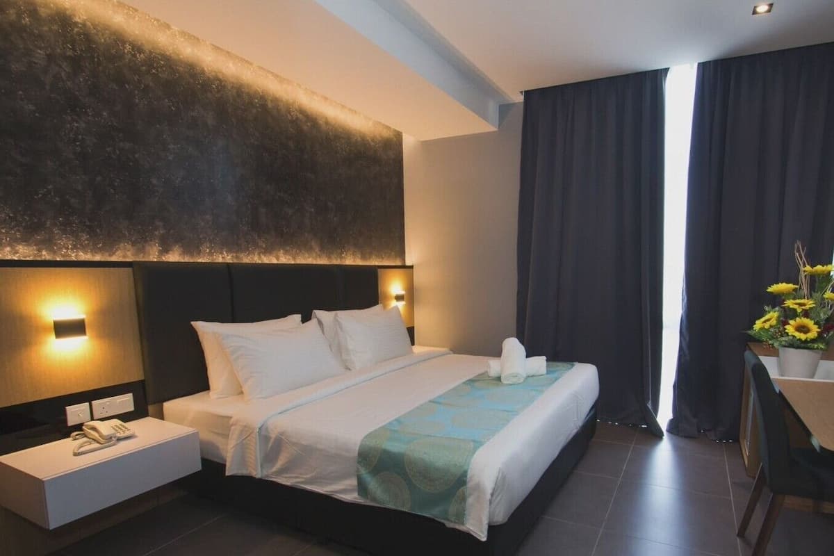 A modern hotel room with a large bed, bedside lamps, a bedside phone, a small table with a vase of flowers, and a view of the outdoors through dark curtains.
