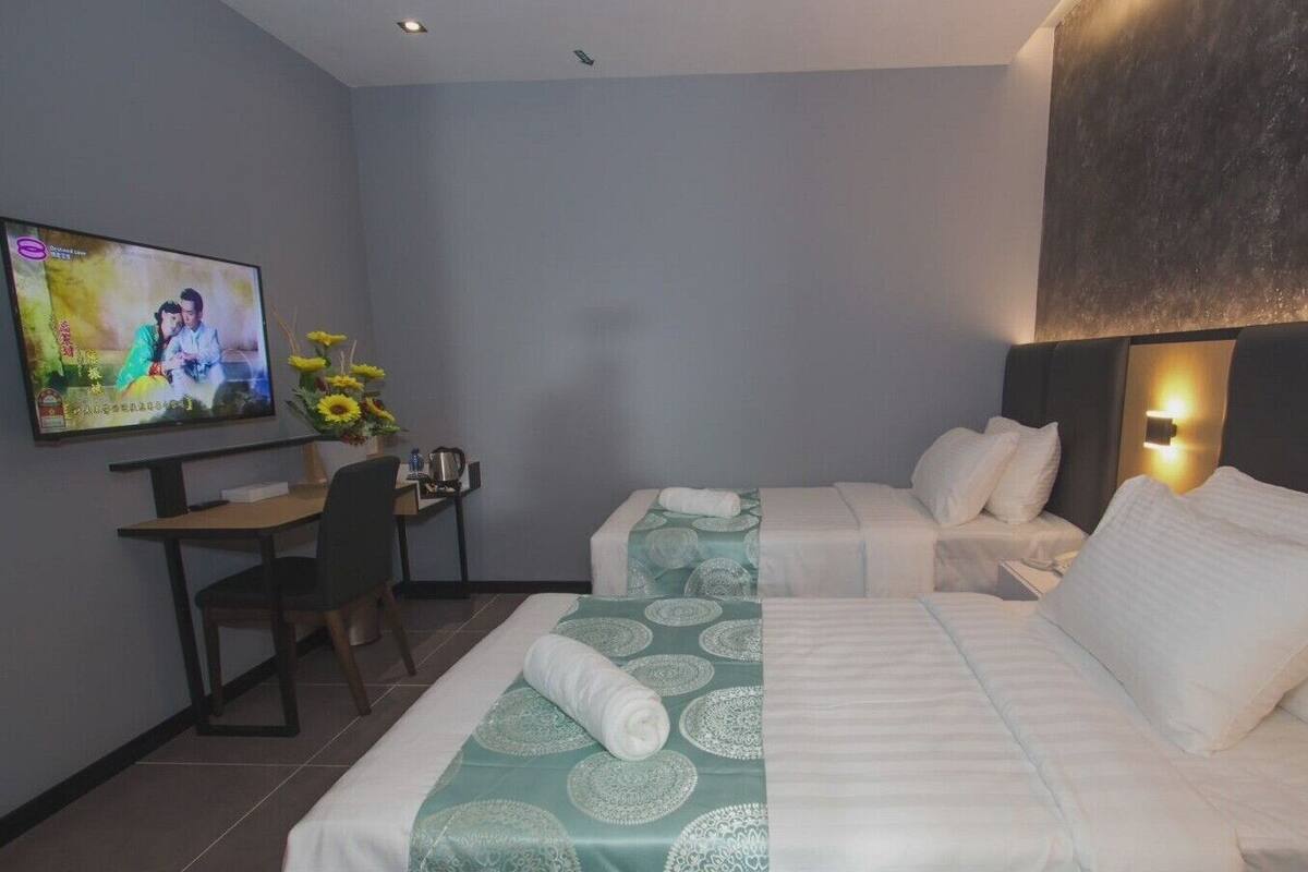 A hotel room with two beds, a desk, a chair, a TV, and a wall-mounted light.