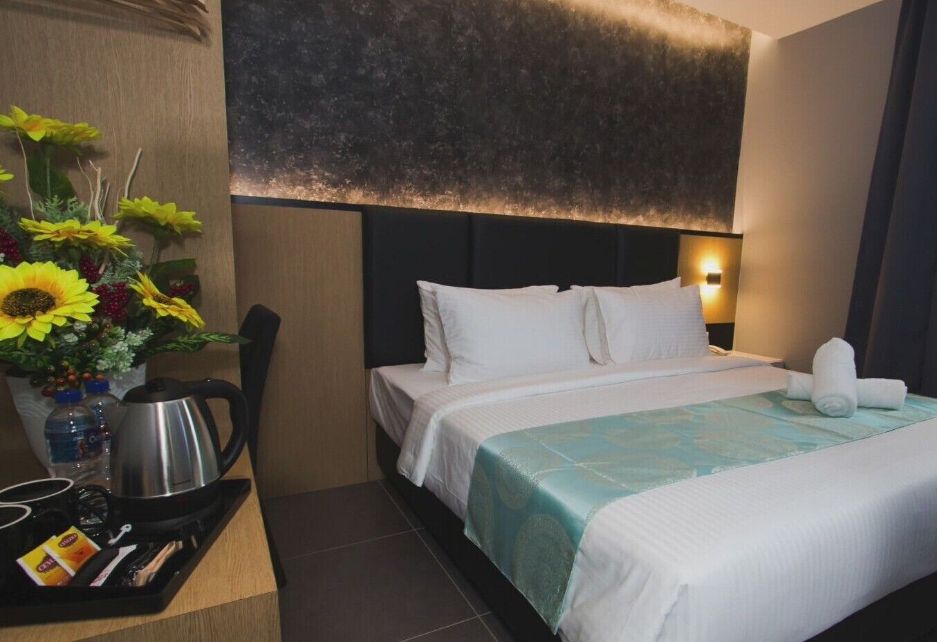 A hotel room with a bed, pillows, a nightstand with a lamp, a vase with flowers, and a tray with a kettle and cups.