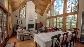 A spacious living room with a fireplace, wooden floors, and large windows offering a view of trees.
