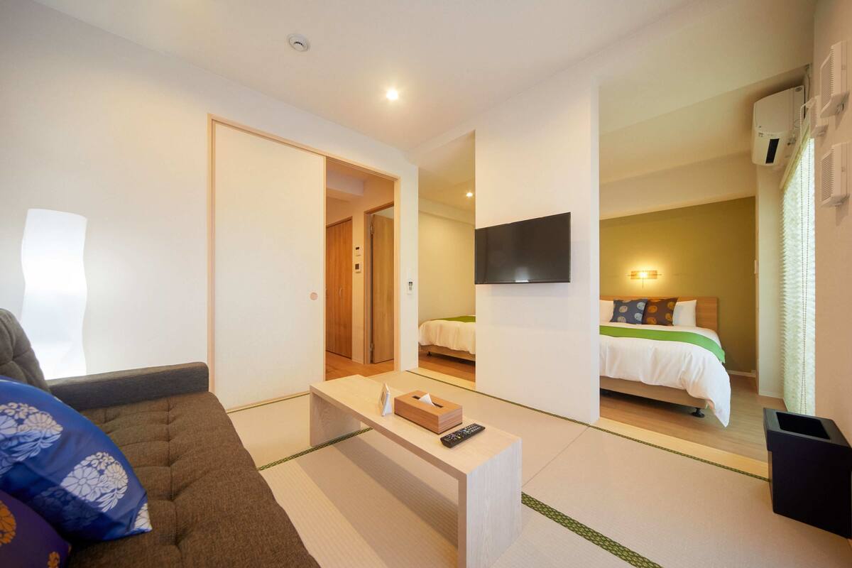 A modern hotel room with a sofa, a small table with a remote control, a bed with a green and white bedspread, and a TV mounted on the wall.