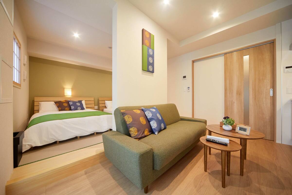 A hotel room with a bed, a sofa, a small table with a clock, and a wall clock.