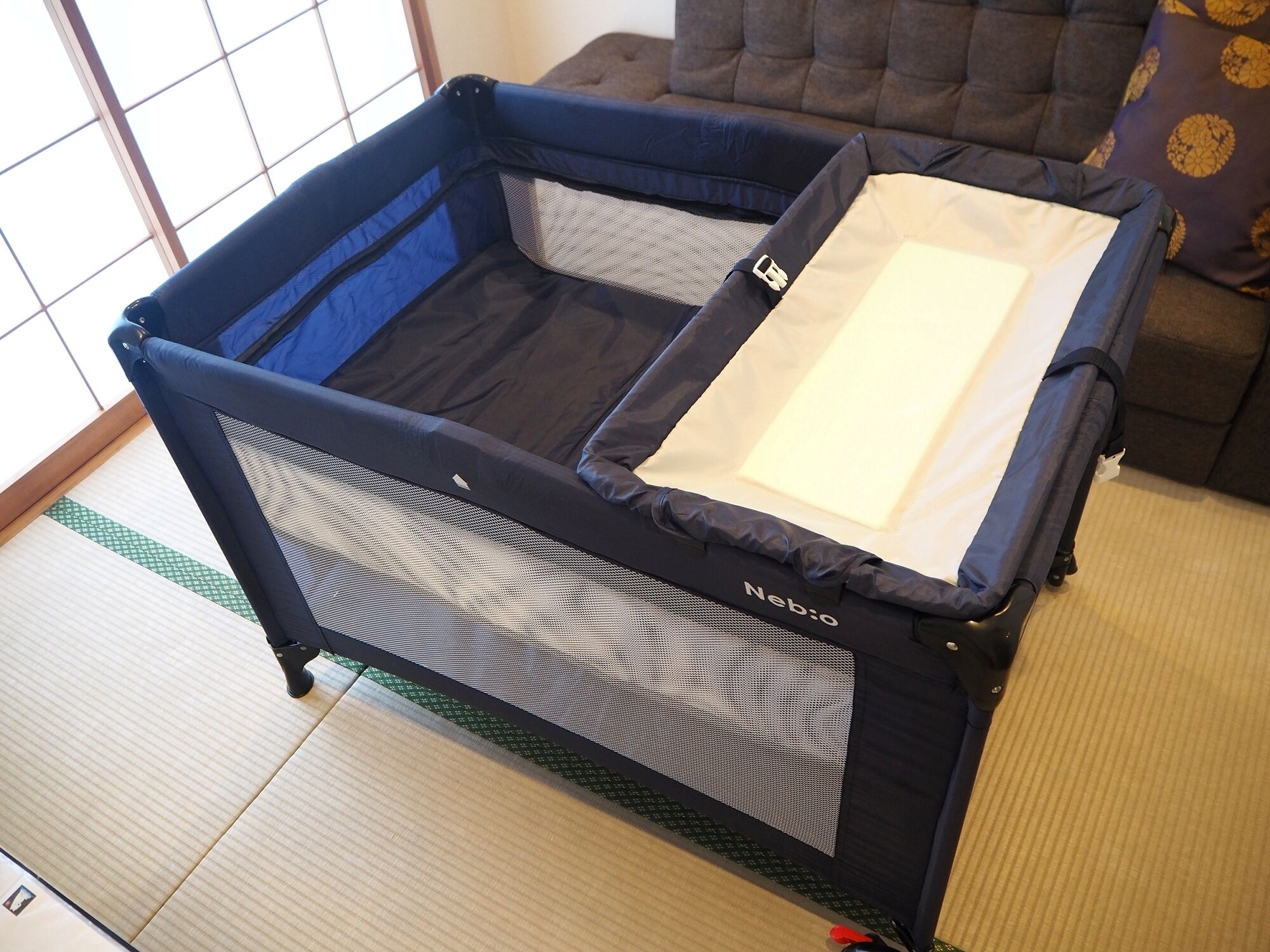 Una habitación con un corralito azul y negro para bebés, piso de esteras de tatami y un sofá con cojines estampados.