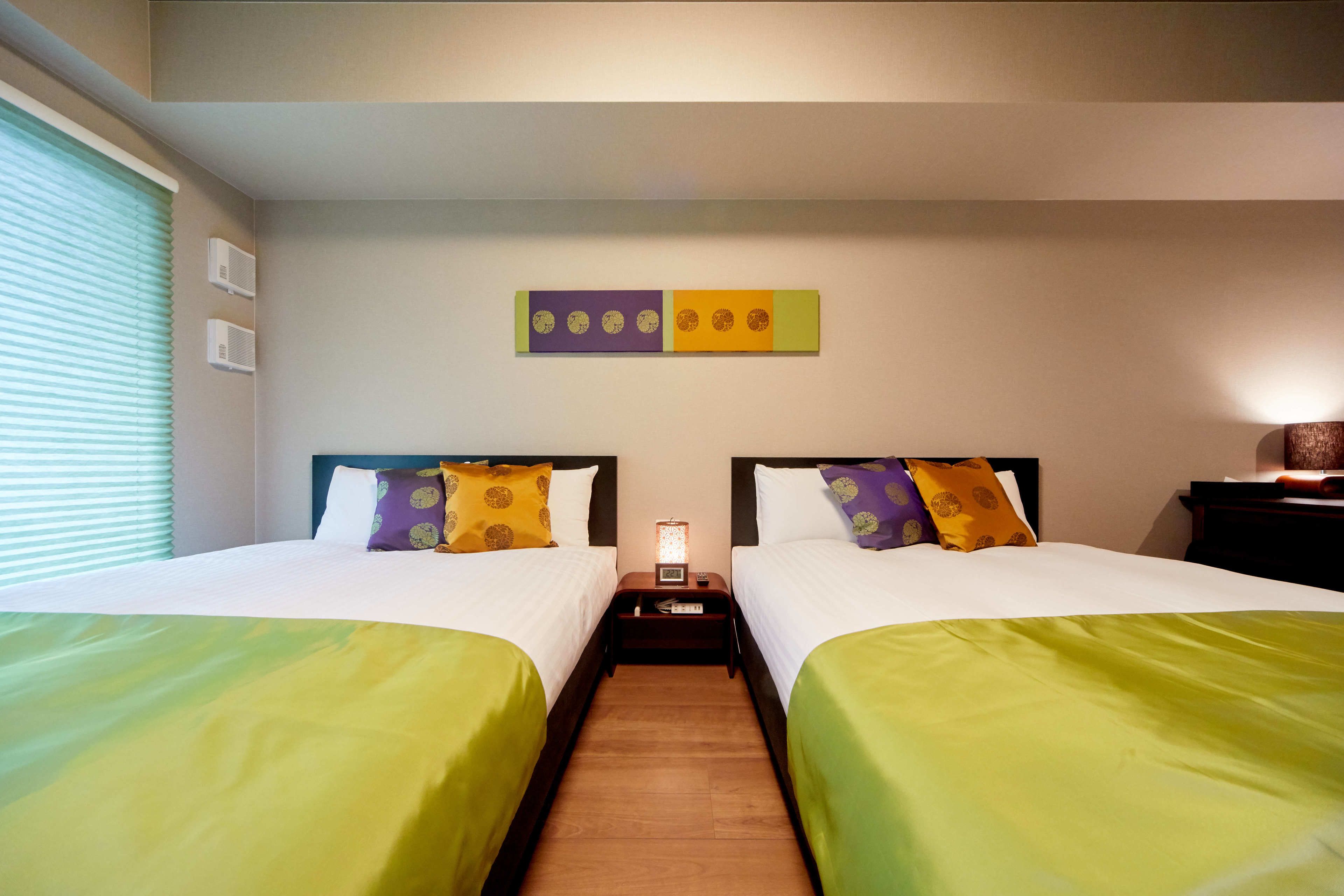 A hotel room with two beds, a nightstand, a wall art piece, and a window with blinds.