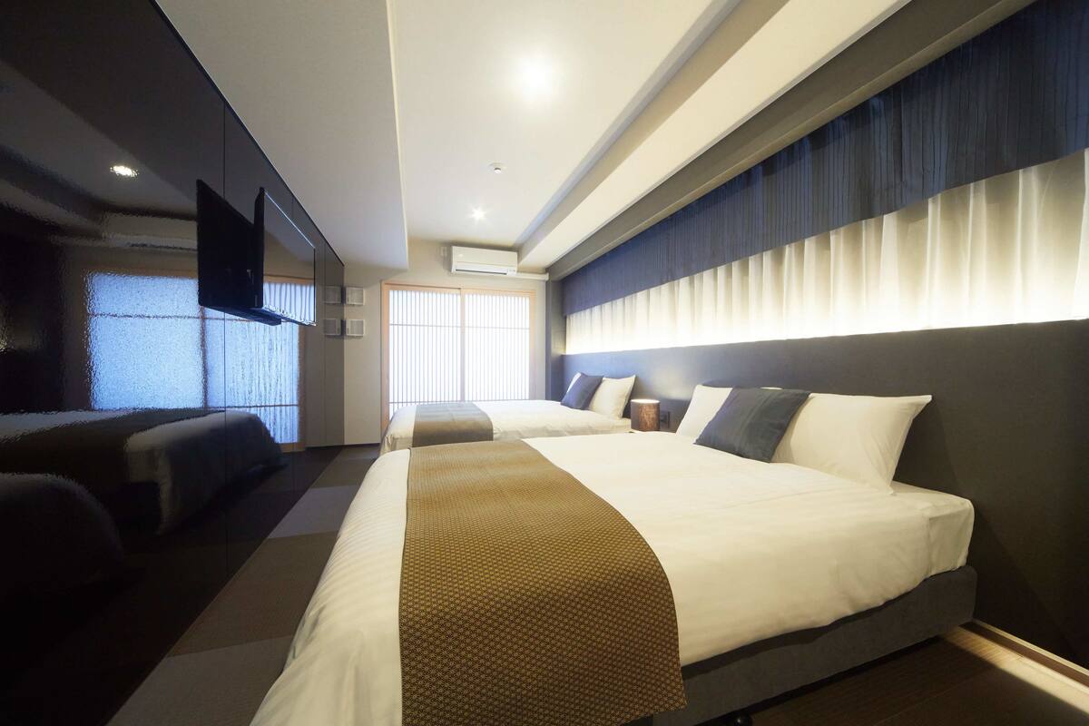 A hotel room with two beds, a flat-screen TV mounted on the wall, and a large window with blinds.