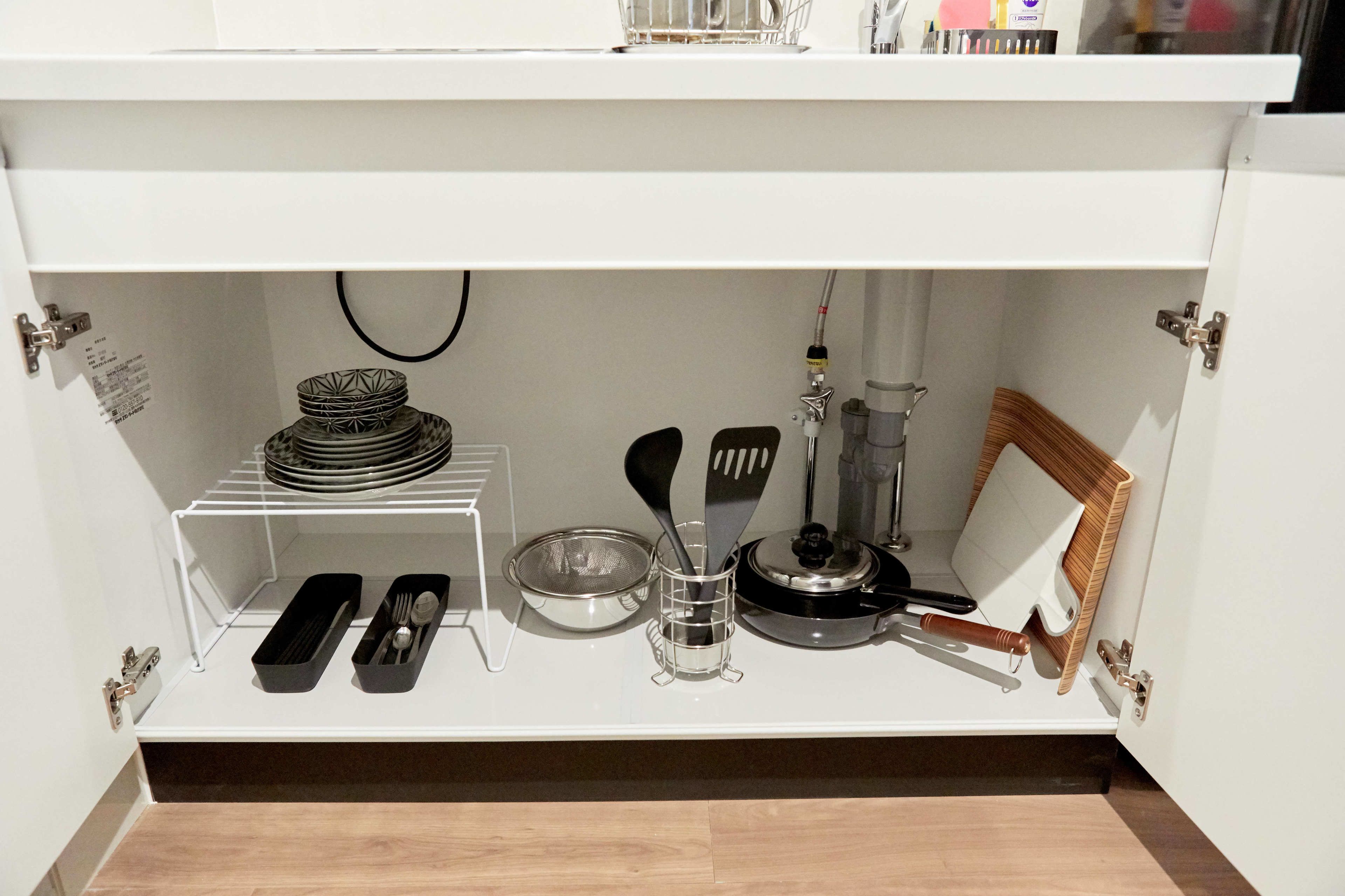 A kitchen cabinet with a shelf containing a stack of plates, a bowl, a colander, a spatula, a whisk, and a small picture frame.