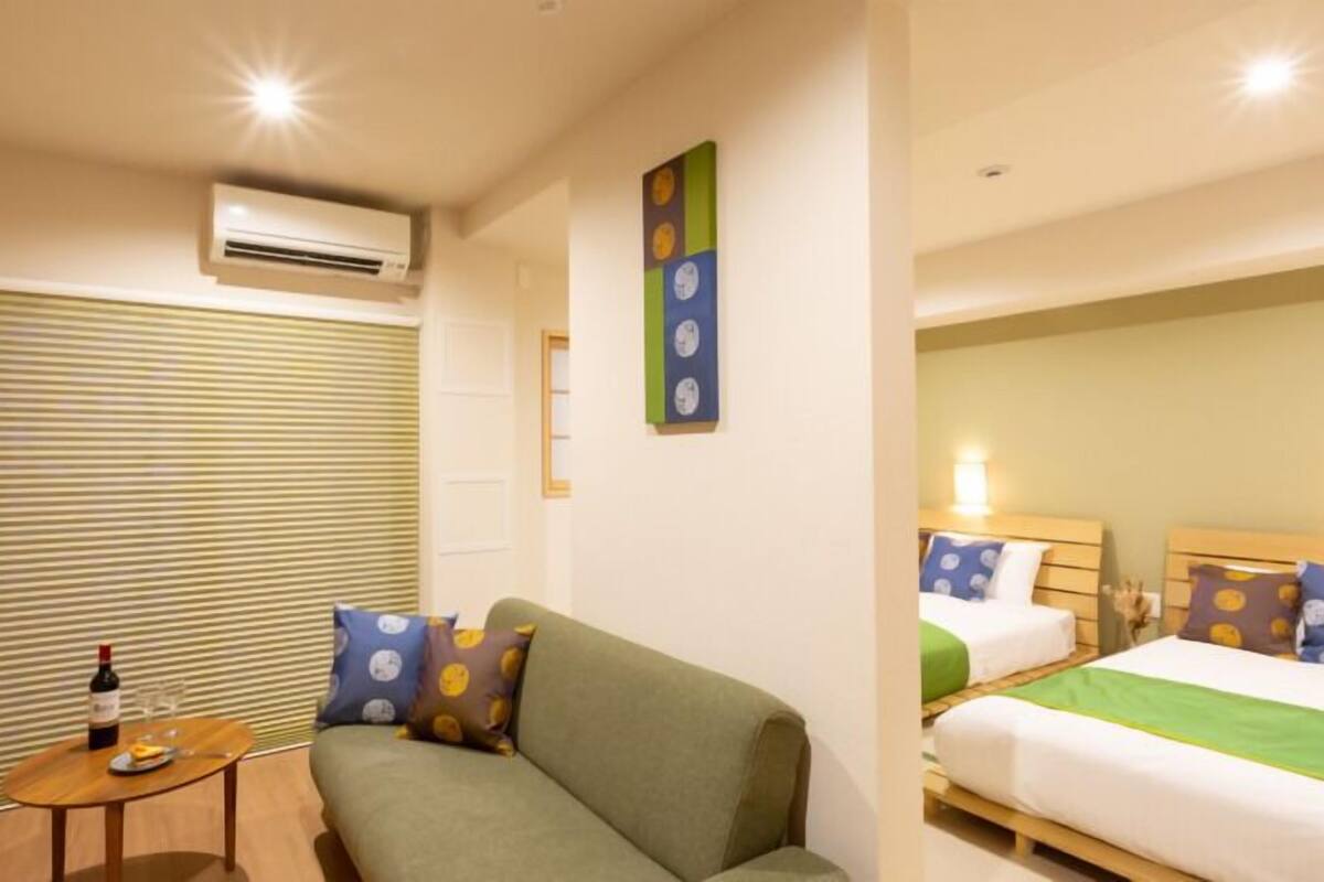 A hotel room with a sofa, a small table with a bottle and glass, a bed with pillows, and an air conditioning unit.